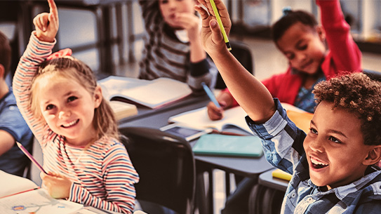 Photo of happy children with their hands up to answer a question, in classroom setting